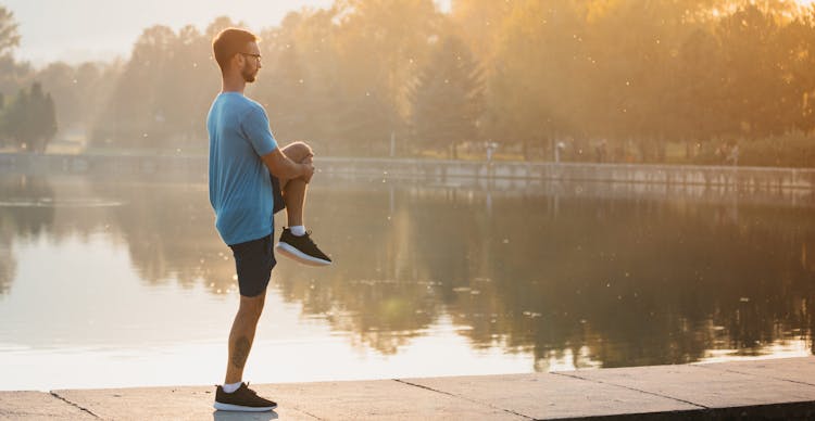 Young man in sportswear stretching by the lake in a city park. R